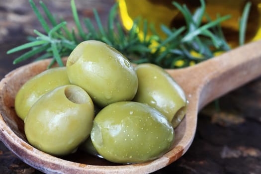 Spanish olives in a wooden spoon with rosemary and olive oil in background on a rustic slate background. Extreme shallow depth of field with selective focus on olives. 