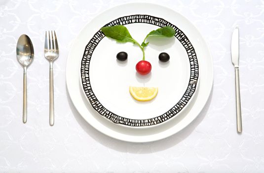 The meal made as a smiling face from olives, radish and lemon slice on a flat plate with a knife, a fork and a spoon
