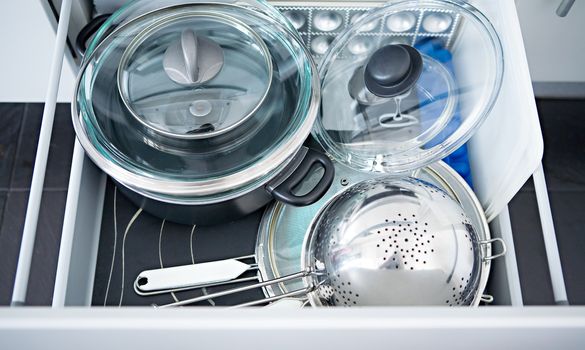 Metal pans with glass covers, a colander and a sieve in an open kitchen box
