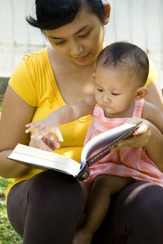 multitasking concept of asian young mother reading a book while babysitting her baby girl outdoor