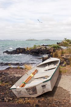 A dinghy rests on the shore of Rangitoto Island in the Hauraki Gulf of New Zealand. Auckland City is in the distance.