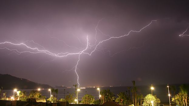 A large multi-fork lightning burst grounding in a town across the bay.  The lights from fishing boats can be seen on the water.  Mountains in the background, palm trees and street lights in the foreground.
