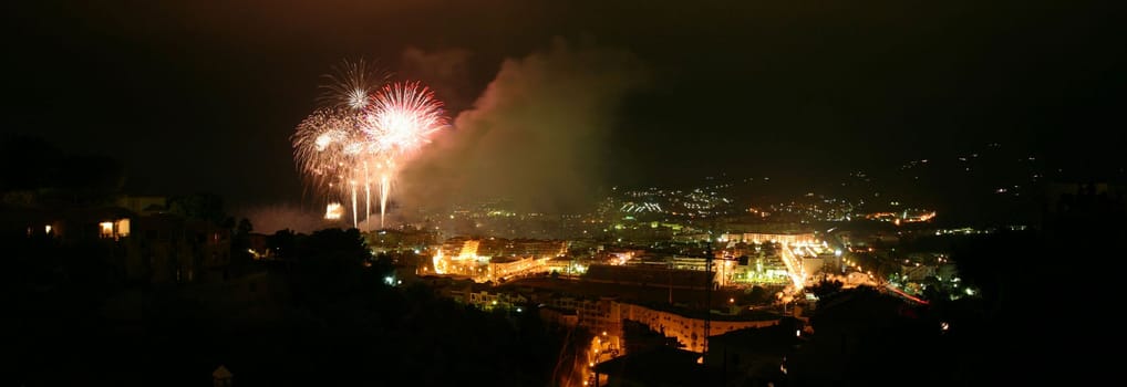 Panoramic view of fireworks at a seaside Spanish fiesta taken from a high viewpoint and overlooking a town.