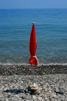 A folded red beach umbrella on an empty rocky beach beside an equally empty sea 