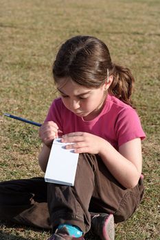 Cute little girl with pigtails writes in the notepad sitting on the grass outdoor