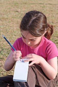 Cute little girl with pigtails writes in the notepad sitting on the grass outdoor