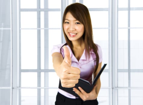 An Asian girl giving thumbs up sign and holding a file in office environment.