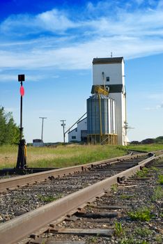 A large grain elevator in the prairies is situated near some empty railroad tracks, shot with a nice blue sky