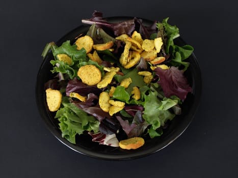 A black plate of salad with crutons and green and purple leaves over a black background.