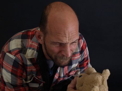 A father holds a childrens stuffed teddy bear, a tear falling down his cheek. Over a black background.