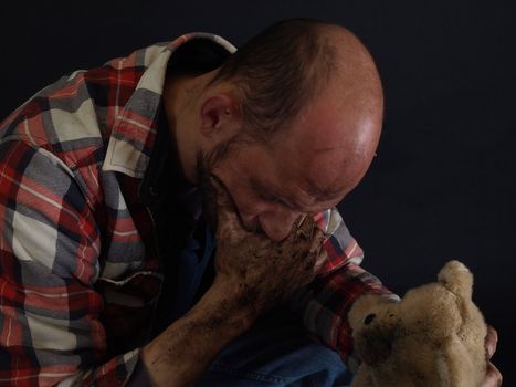 A dirt covered man holds an orphaned dirty stuffed teddy bear in one hand, his jaw in another. Over a black background.