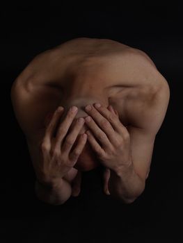 A nude male cowers, his head covered with his hands, over a black background.
