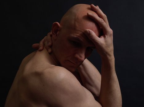 A nude bald male in the shadows over a black background.