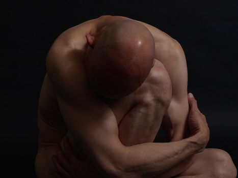 A clean shaven nude male sits in darkness, crumpled up. Over a black background.