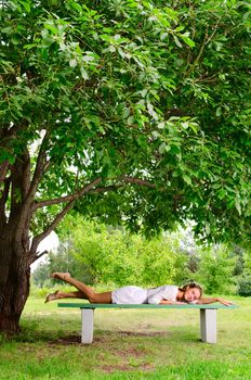 Pretty barefooted woman with  is laying on a bench under the green tree. She is slightly smiling and looking at the camera