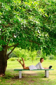 Pretty barefooted woman is laying on a bench under the green tree. She is working with small notebook. She is looking at the camera and slightly smiling