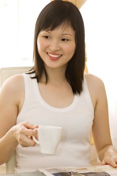 A beautiful young Asian woman drinking tea and reading newspaper. 
