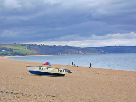 Small fishing boat moored on a beach in South Devon in England with two men rod fishing in the distance.