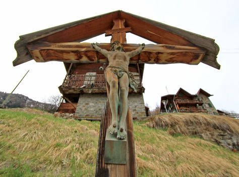 Brown wood cross with Jesus sculpture on it in front of chalet jouses in the mountain