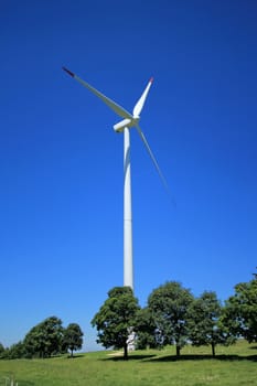 Red and white wind turbine surrounded with trees and green grass by beautiful weather and deep blue sky