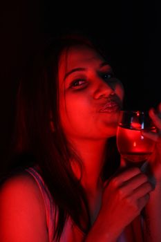 A beautiful Indian woman kissing a glass of wine, in red party lights.