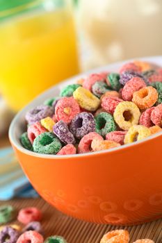 Colorful cereal loops with different fruit flavour in orange bowl with orange juice and milk in the back (Selective Focus, Focus one third into the bowl)