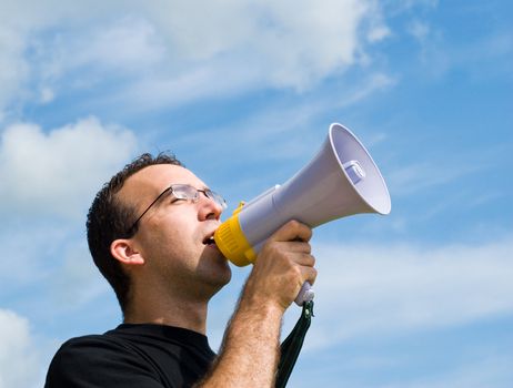 A young man wearing glasses is talking into a megaphone outside with blue sky behind him