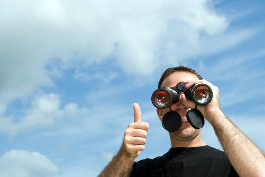 A young man gives a thumbs up as he holds a set of binoculars to his eyes, with clouds and blue sky behind him