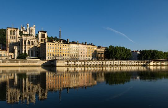 Image shows a cityscape of Lyon, France taken from an angle showing the Notre Dame de Fourviere basilica as well as buildings on the bank of the Saone river