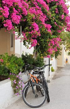 Scene from a narrow alley from a Greek island village, showing two bicycles leaning against a house covered in colorful flowers
