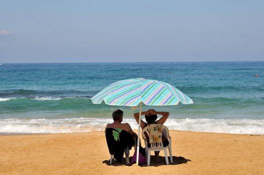 couple sitting on chairs under an umbrella on the beach
