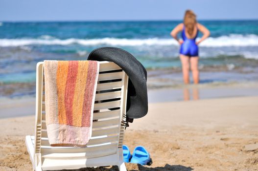
	
A hat, a towel hanging on a chair on the beach by the sea, and in the distance a woman 