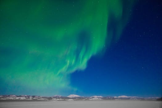 Northern Lights (Aurora borealis) over moon lit snowscape of frozen lake and forested hills.