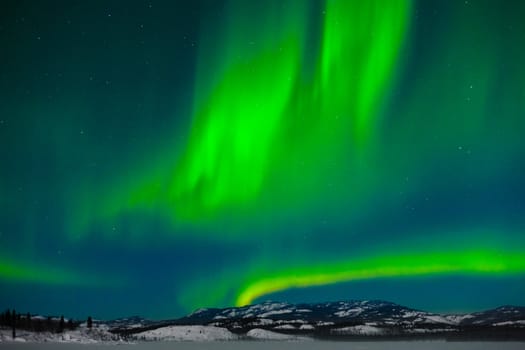 Northern Lights (Aurora borealis) over moon lit snowscape of frozen lake and forested hills.