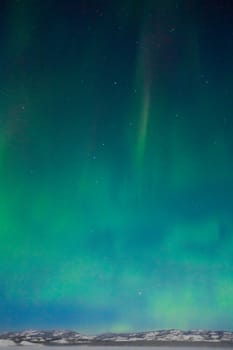 Northern Lights (Aurora borealis) over moon lit snowscape of frozen lake and forested hills.