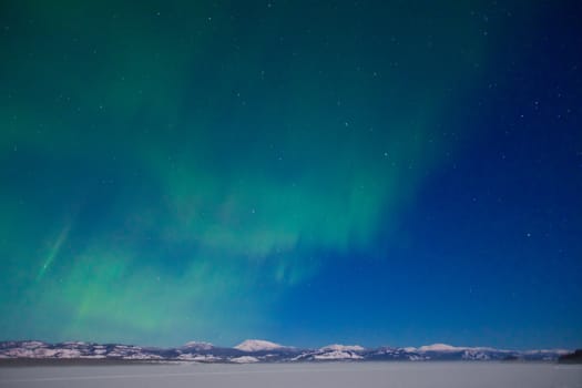 Northern Lights (Aurora borealis) over moon lit snowscape of frozen lake and forested hills.