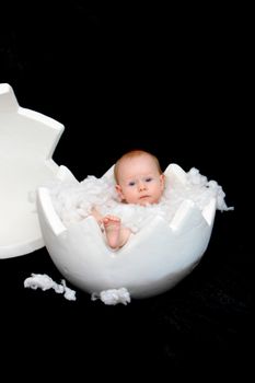 A baby sits in a large egg-shell.The background is black