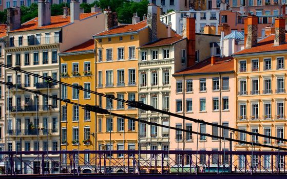 Image shows a series of colorful buildings in the city of Lyon, France, washed in the warm afternoon sunlight. One of the famous Lyon hanging bridges is seen in the foreground.