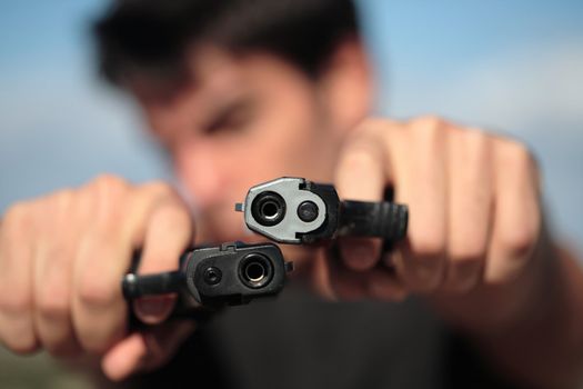 A young, robust man, in his 20's with dark hair pointing 2 pistols to the camera.