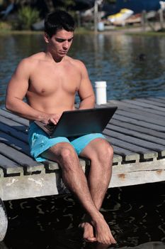 A young man using a laptop on a pier, by the lake, on a warm day, wearing no shirt. 
