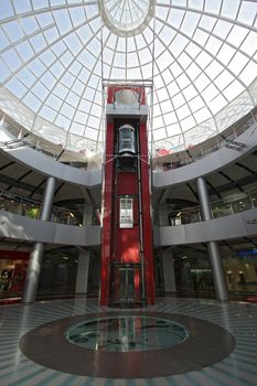 Huge hall of modern shopping centre with the glass lift, hours on a floor and a transparent glass dome