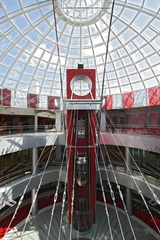 Huge hall of modern shopping centre with the glass lift, hours on a floor and a transparent glass dome