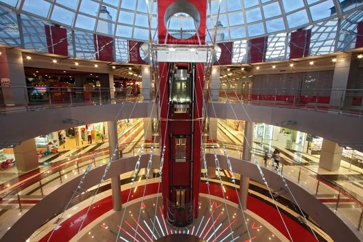 Huge hall of modern shopping centre with the glass lift, hours on a floor and a transparent glass dome