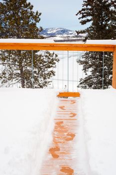 Footsteps of person trying to escape winter by jumping over railing of snow covered wooden deck.