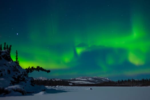 Northern Lights (Aurora borealis) over moon lit snowscape of frozen lake and forested hills.