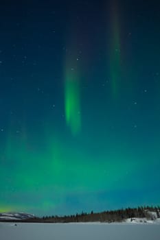 Northern Lights (Aurora borealis) over moon lit snowscape of frozen lake and forested hills.