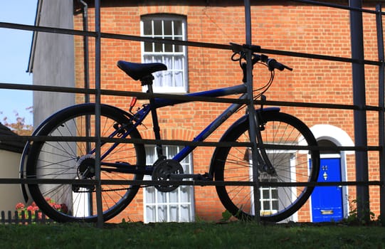 A bike resting against a set of railings with a red bricked terraced house with a blue door visible to the background.
