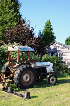 A farm tractor in a field located in a grassed field in rural Wiltshire, UK england. Set against a backdrop of a wooden outbuilding, hedgerows and trees.