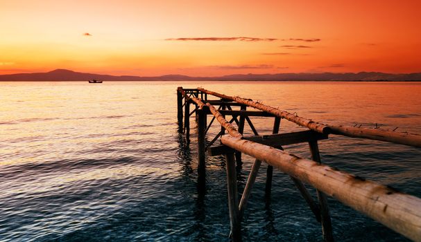 A primitive wooden pier, photographed during a glorious sunset,  extends into the sea and leads the eye to a fishing boat in the distance.