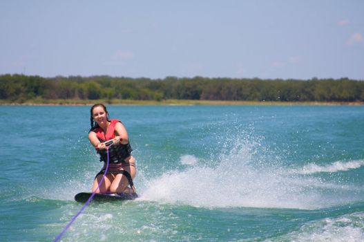 A pretty 20 something girl smiles while kneeboarding on a blue lake. 